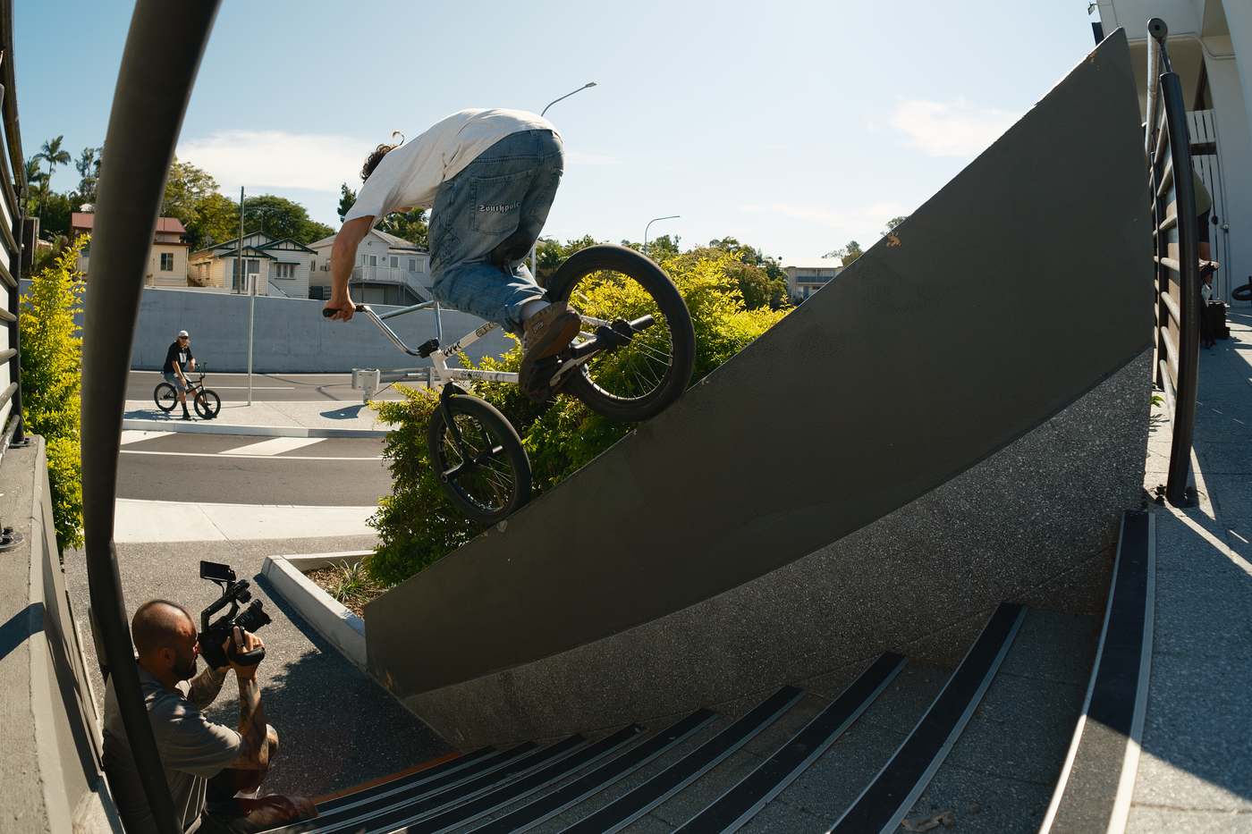 Kai riding along a steep angled concrete wall above a stairset, a filmer crouched below, shot on a fisheye.
