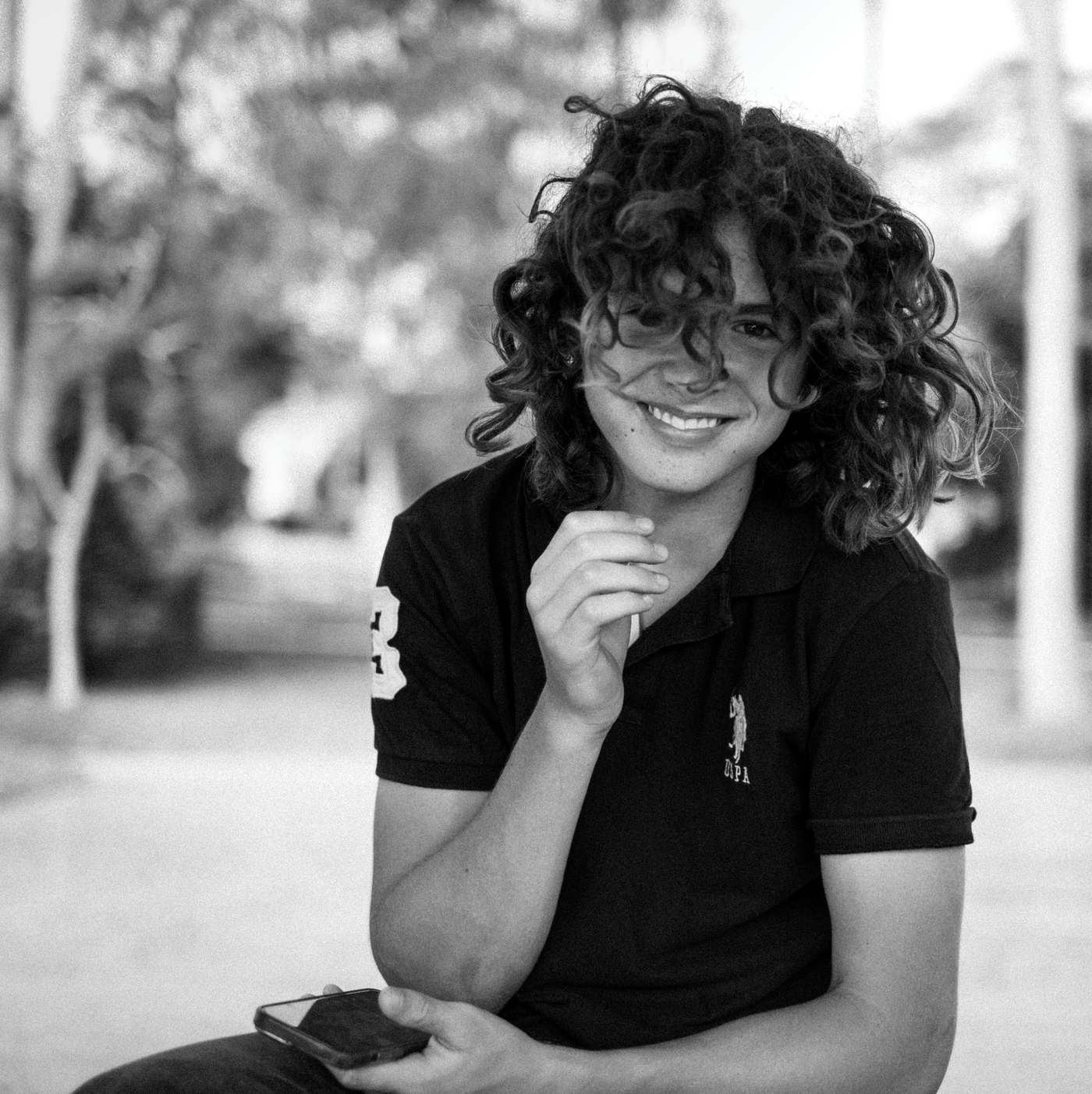 Kai Hayes with long curly hair, grinning, phone in hand, in black and white.