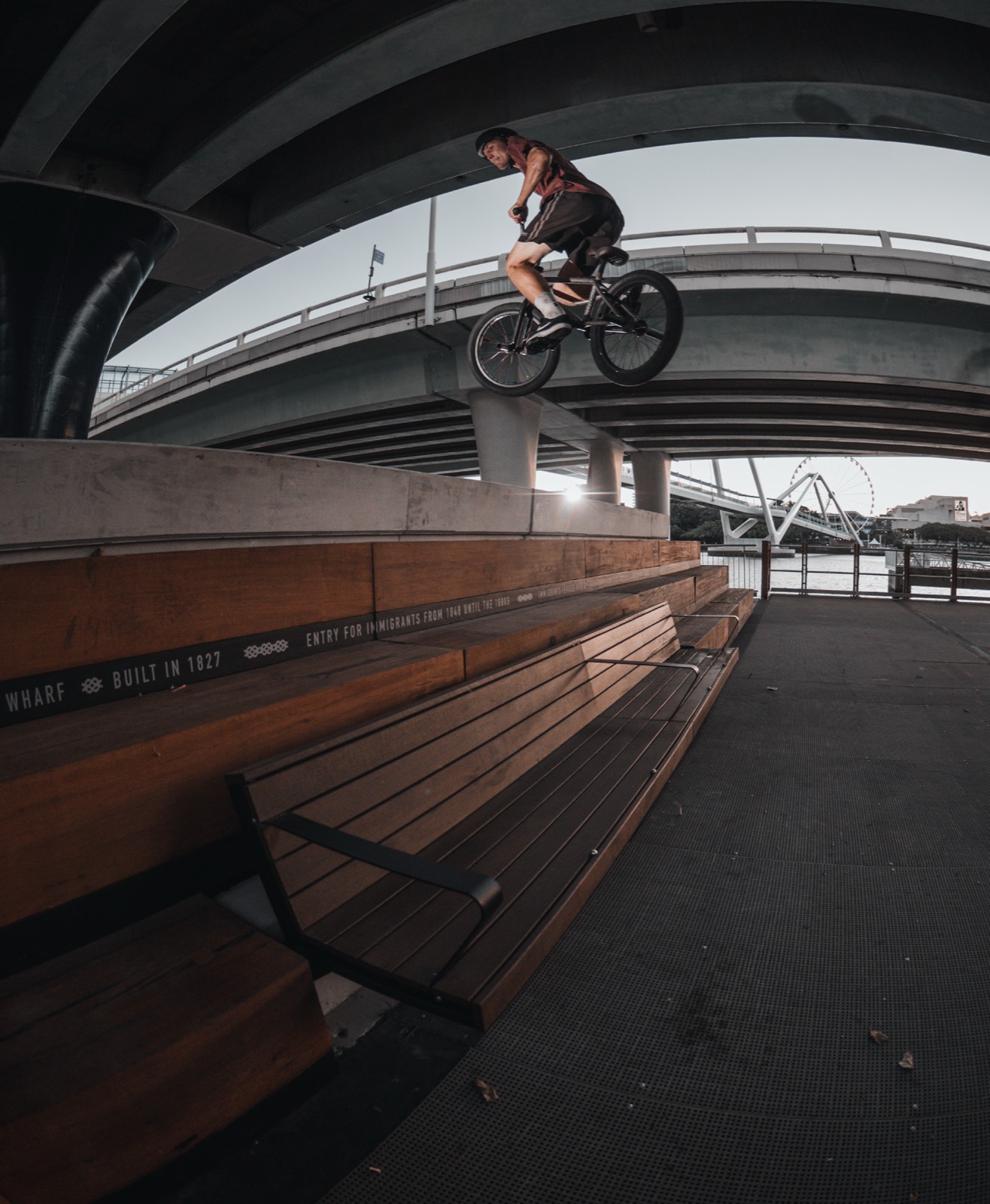 Nathan Smith mid-air in a 360 off a riverside bench under a Brisbane bridge, shot on a fisheye.